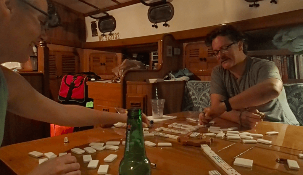 Domino game stretched out on the salon table with Margaret and Felipe reaching for tiles. A beer bottle and empty rum drink cup are in the frame.