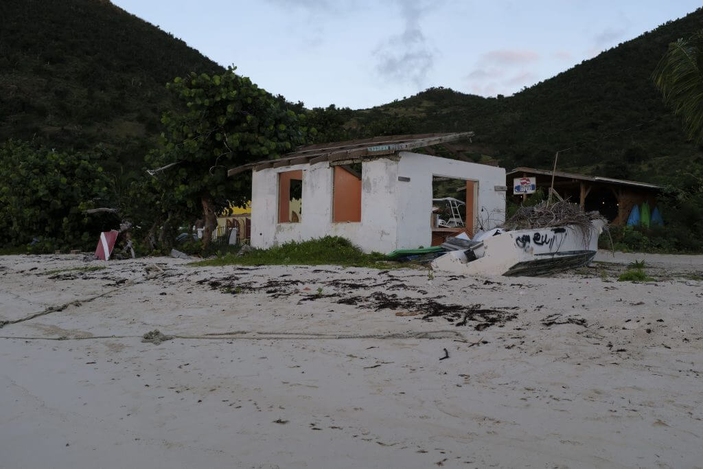 white cement building with no doors and windows on the beach with a banged up motor boat alongside