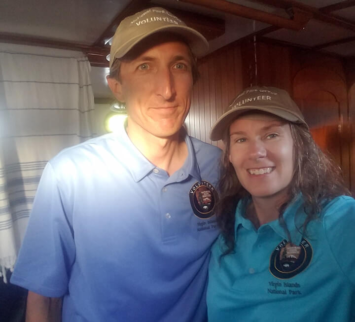 A man and a woman in a sailboat wearing Virgin Island Nation Park Volunteer uniform polo shirts and hats.