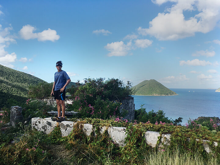 Man standing in ruins of Murphy House on St John Island, USVI. Man is wearing blue shirt, visor, sunglasses and black shorts. Green vegetation and pink flowers abound.